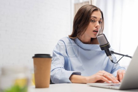 Brunette broadcaster talking at microphone and using blurred laptop near coffee to go in paper cup on table during stream in podcast studio 