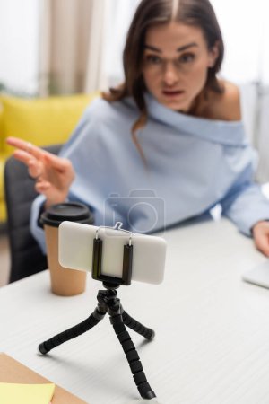Blurred brunette broadcaster pointing with finger and talking while looking at smartphone on tripod near coffee to go on table during stream in podcast studio 