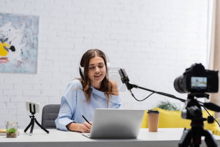 Uśmiechnięta brunetka podcaster w słuchawkach za pomocą laptopa i trzymając długopis w pobliżu mikrofonu i aparatu cyfrowego w pobliżu stołu podczas strumienia w studio 