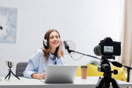 Digital camera near blurred brunette broadcaster in headphones talking during stream near laptop and smartphone on table in podcast studio 