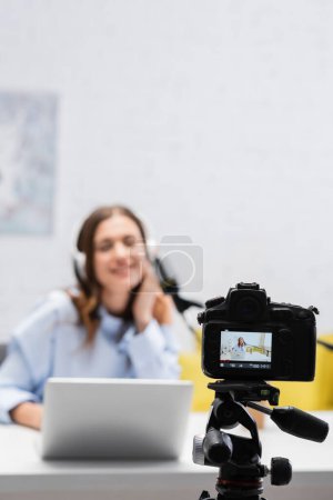Digital camera on tripod near blurred brunette broadcaster in headphones using laptop near microphone during stream in podcast studio 