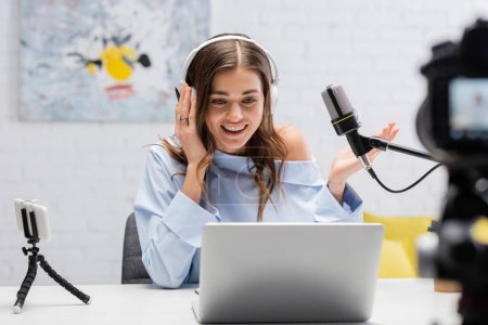 Beztroski brunetka podcaster w słuchawki rozmowy i patrząc na laptopa w pobliżu mikrofonu i rozmyty aparat cyfrowy podczas strumienia w studio 