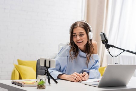 Lächelnde brünette Podcasterin in drahtlosen Kopfhörern mit Smartphone auf Stativ und Laptop in der Nähe von Mikrofon und Notebooks auf dem Tisch während des Streams im Studio 