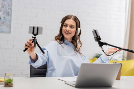 Brunette podcaster in wireless headphones smiling during stream and holding smartphone on tripod near microphone and laptop in studio 