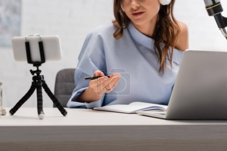 Photo for Cropped view of brunette podcaster in blur blouse talking during stream near laptop, smartphone on tripod and microphone during stream in studio - Royalty Free Image
