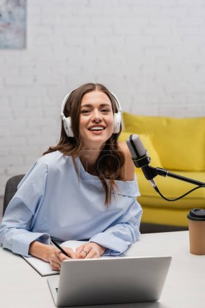 morena positiva podcaster en auriculares mirando a la cámara mientras escribe en el portátil cerca de la computadora portátil y el micrófono durante la corriente en el estudio 