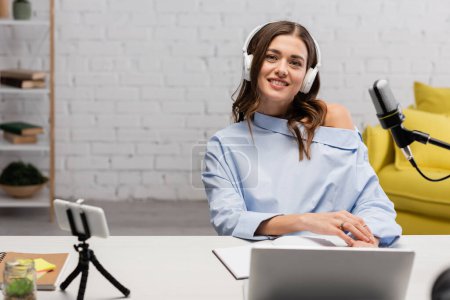 Uśmiechnięty brunetka nadawca w niebieskiej bluzce i słuchawki bezprzewodowe patrząc na kamerę w pobliżu mikrofonu, gadżety i notebooki w studio 