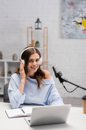 Wesoła brunetka podcaster w słuchawkach patrząc na aparat w pobliżu urządzeń, notebook, filiżanka papieru z kawą na wynos i mikrofon podczas strumienia w studio podcast 