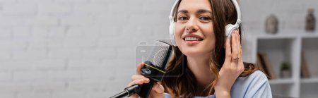 Brunette podcaster smiling and touching wireless headphones holding microphone and looking at camera during stream in podcast in studio, banner 