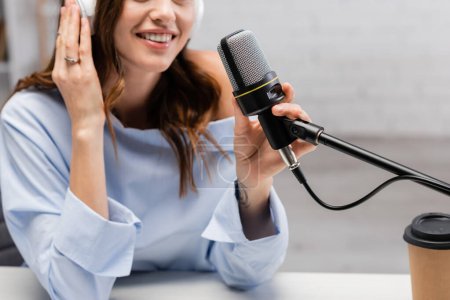 Ausgeschnittene Ansicht eines verschwommenen und positiven Podcast-Moderators in drahtlosen Kopfhörern, der in der Nähe von Mikrofon und Kaffee zum Mitnehmen in Pappbecher auf dem Tisch im Studio spricht 