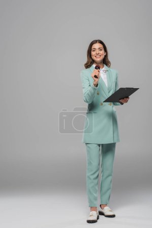 Full length of smiling and brunette event host in blue suit holding microphone and clipboard while looking at camera and standing on grey background 