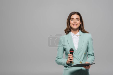 Unbekümmerte und brünette Moderatorin in blauer Festkleidung, die Mikrofon und Klemmbrett in die Kamera hält und isoliert auf grau mit Kopierraum steht