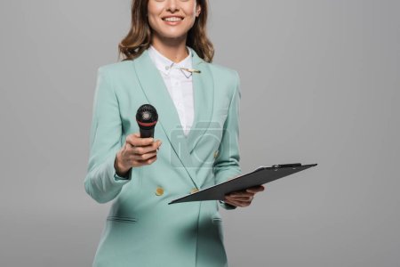 Cropped view of smiling and brunette event host in blue formal wear holding wireless microphone and clipboard during celebration isolated on grey  