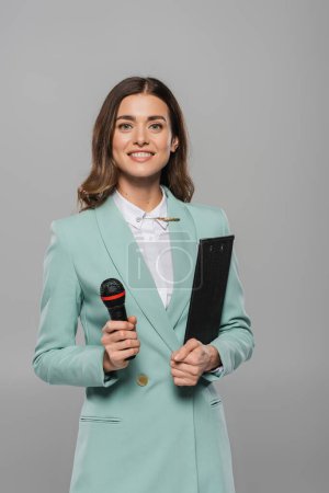 Portrait of positive brunette host of event in blue suit holding microphone and clipboard during party while looking at camera and standing isolated on grey  