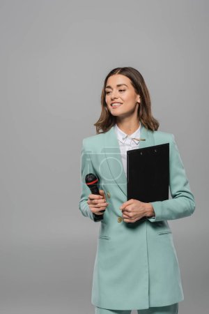 Cheerful brunette host of event in blue jacket and shirt holding microphone and clipboard while looking away during celebration isolated on grey  