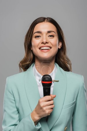 Portrait of smiling brunette event host in blue formal wear talking at wireless microphone and looking at camera while standing isolated on grey  