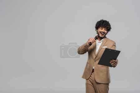 Photo for Young and overjoyed indian event host in suit talking at microphone and looking at clipboard during party while standing isolated on grey with copy space - Royalty Free Image