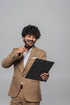 Photo for Young indian host of event in formal wear smiling while talking at microphone and holding clipboard during party and standing isolated on grey - Royalty Free Image