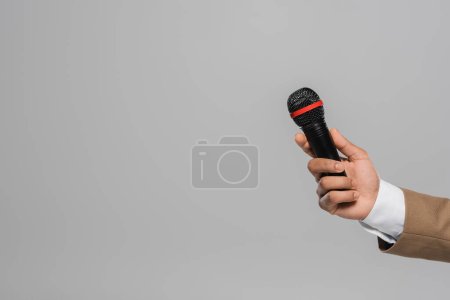 Vista recortada de la mano del anfitrión del evento en desgaste formal marrón que sostiene el micrófono inalámbrico mientras está de pie aislado en gris con espacio de copia