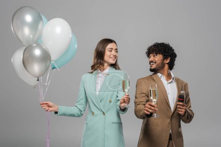 Téléchargez les photos : Des hôtes interracial positifs en tenue formelle avec microphone et ballons festifs tenant des verres de champagne et se regardant isolés sur gris - en image libre de droit