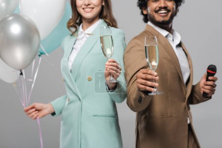 Foto de Vista recortada de sonrientes anfitriones de eventos interracial en ropa formal sosteniendo micrófono, globos festivos y copas de champán mientras están aislados en gris - Imagen libre de derechos