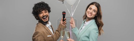 Téléchargez les photos : Des hôtes interraciaux positifs en costumes tenant des ballons festifs, un microphone et des verres de champagne tout en restant ensemble isolés sur une bannière grise - en image libre de droit