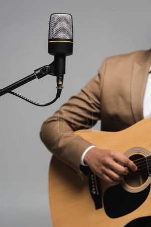 Ausgeschnittene Ansicht des Mikrofons auf dem Stativ in der Nähe verschwommener junger Moderator spielt Akustikgitarre während des Auftritts und Party isoliert auf grau  