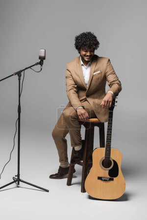Foto de Joven y sonriente anfitrión del evento indio en traje mirando la guitarra acústica mientras está sentado en la silla cerca del micrófono durante la actuación sobre fondo gris - Imagen libre de derechos
