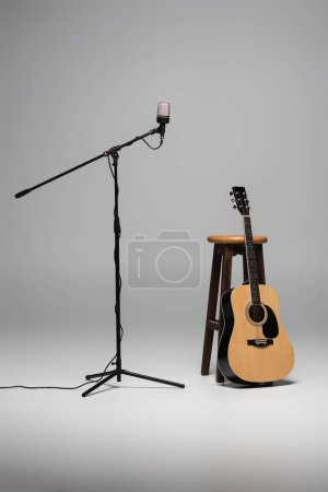 Microphone sur support métallique et guitare acoustique près d'une chaise brune en bois sur fond gris 