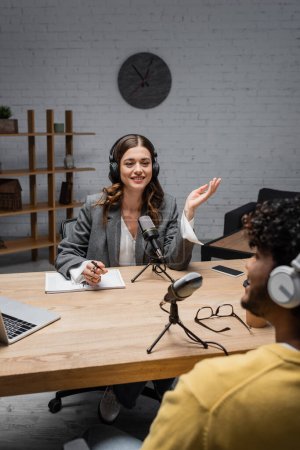 Smiling podcast host in wireless headphones talking to blurred indian guest near notebook, pen, glasses, microphones and devices with copy space during stream in studio 