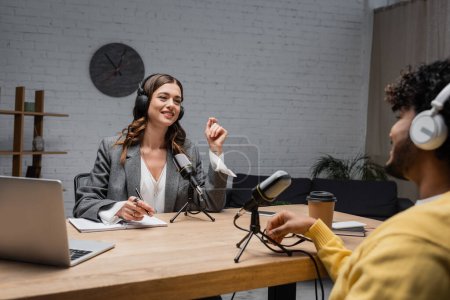 positive female podcast host in wireless headphones talking to blurred indian guest while sitting with pen near notebook, devices, microphone and coffee to go during stream in studio 
