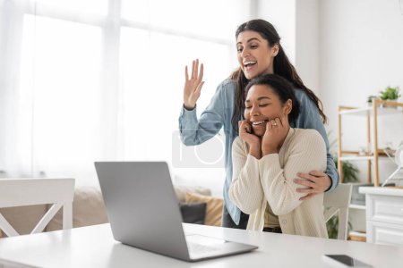 glückliche lesbische Frau winkt bei Videoanruf in der Nähe ihrer multirassischen Freundin mit Verlobungsring am Finger