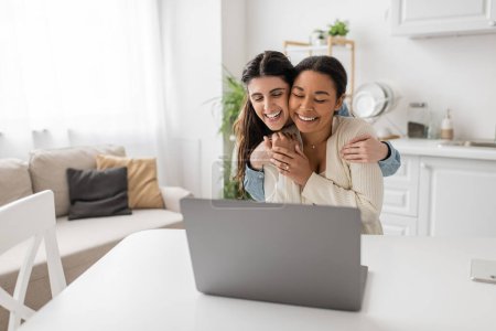 happy lesbian woman hugging multiracial girlfriend with engagement ring on finger near laptop during video call 