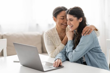 joyful interracial lesbian couple with closed eyes hugging near devices on table 