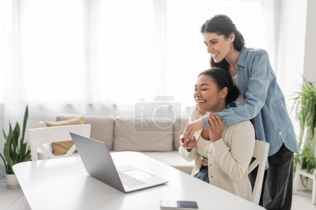 joyful multiethnic lesbian couple showing engagement ring during video call on laptop