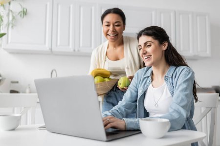 Photo pour Heureuse femme multiraciale tenant bol avec des fruits près de petite amie gaie travaillant sur ordinateur portable à la maison - image libre de droit
