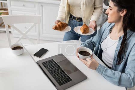 woman holding plates with baked croissants near cheerful girlfriend working on laptop from home 