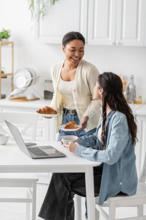 multiracial woman holding plates with baked croissants near happy girlfriend working on laptop from home 