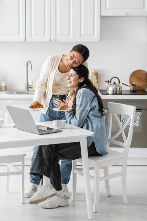 happy multiracial woman holding plates with croissants near girlfriend working on laptop from home 