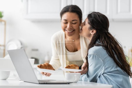 happy lesbian woman kissing cheek of cheerful multiracial girlfriend with croissants  