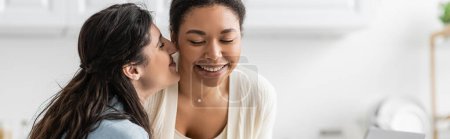 happy lesbian woman kissing cheek of cheerful multiracial girlfriend, banner 