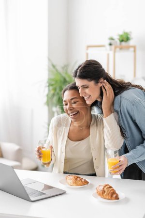 cheerful interracial lesbian couple looking at laptop while holding glasses of orange juice 