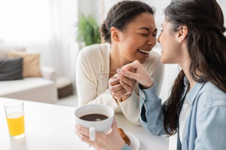 Lesbische Frau mit geschlossenen Augen lacht, während sie beim Frühstück die Hand ihres Partners hält 