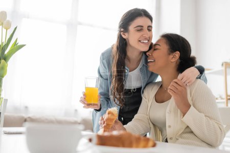 Photo pour Gaie lesbienne femme tenant verre de jus d'orange et étreignant petite amie multiraciale pendant le petit déjeuner dans la cuisine - image libre de droit