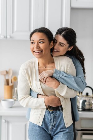 cheerful lesbian woman hugging positive multiracial girlfriend in modern kitchen 