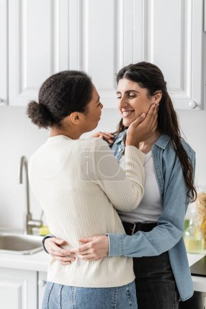 pleased lesbian woman hugging with multiracial girlfriend in modern kitchen 