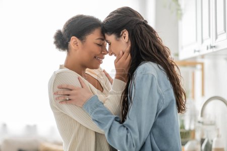 lockige multirassische Frau umarmt fröhliche Freundin mit geschlossenen Augen zu Hause 