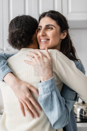 happy lesbian woman with wedding ring on finger smiling and hugging multiracial girlfriend  