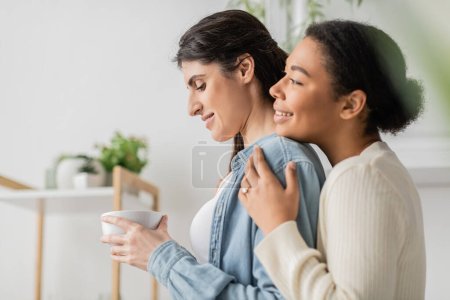 overjoyed multiracial woman with engagement ring on finger hugging girlfriend holding cup of coffee 