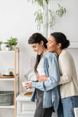 overjoyed multiracial woman with engagement ring on finger hugging happy girlfriend holding cup of coffee 